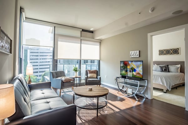 Modern apartment living room with floor-to-ceiling windows overlooking city skyline, gray sofa, armchairs, round coffee table, and bedroom visible through open
