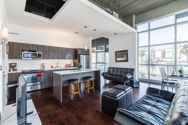 Open-plan kitchen and living room with dark wood cabinetry, stainless steel appliances, island seating, black leather furniture, and floor-to-ceiling windows