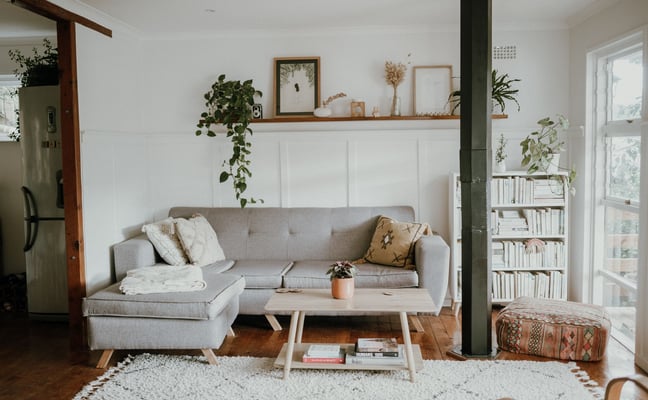 Bright living room with gray sectional sofa, wooden coffee table, white bookshelf, potted plants, and wood beam ceiling